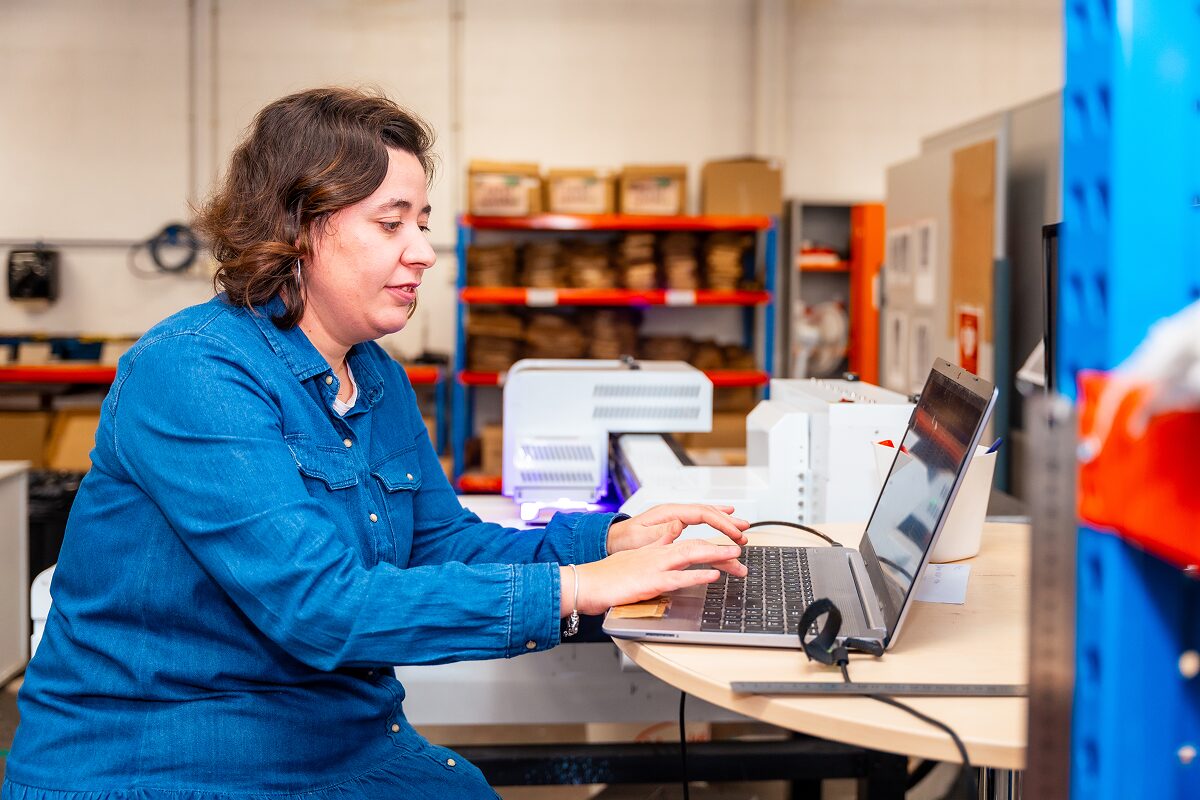 A woman seated at a workbench types on a laptop in an industrial workspace with shelves of supplies behind her.