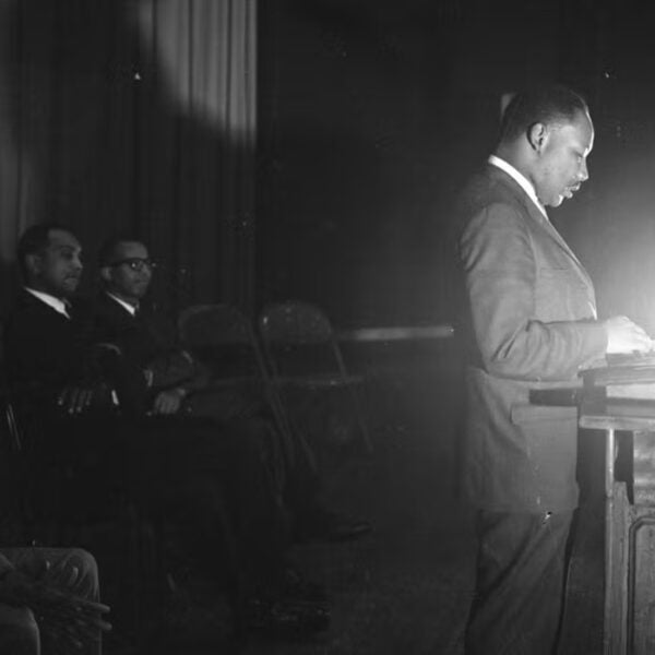 * Black-and-white photo of Martin Luther King Jr. standing at a podium, reading under a bright stage light, with men seated behind him.