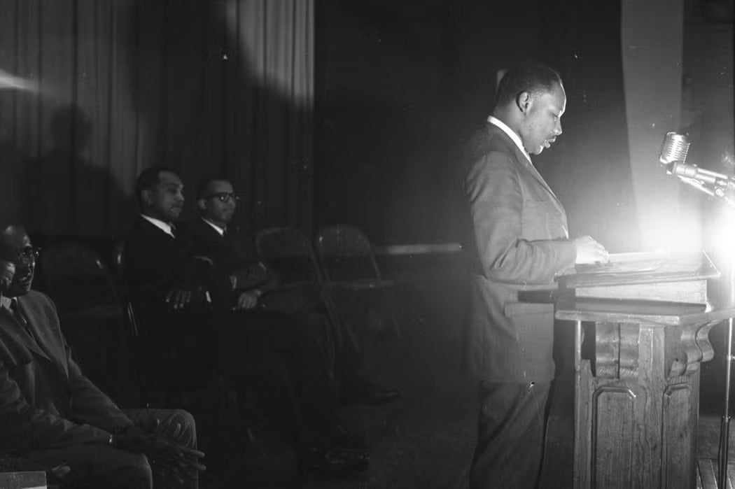 Black-and-white photo of Martin Luther King Jr. standing at a podium, reading under a bright stage light, with men seated behind him.