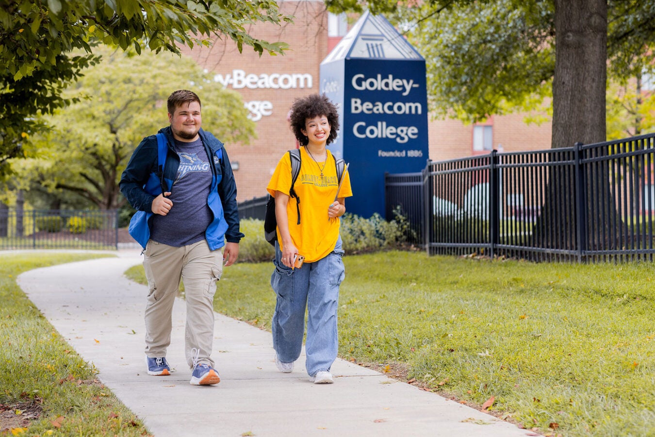 Two college students walking along a tree-lined campus path near a Goldey-Beacom College sign, carrying backpacks.