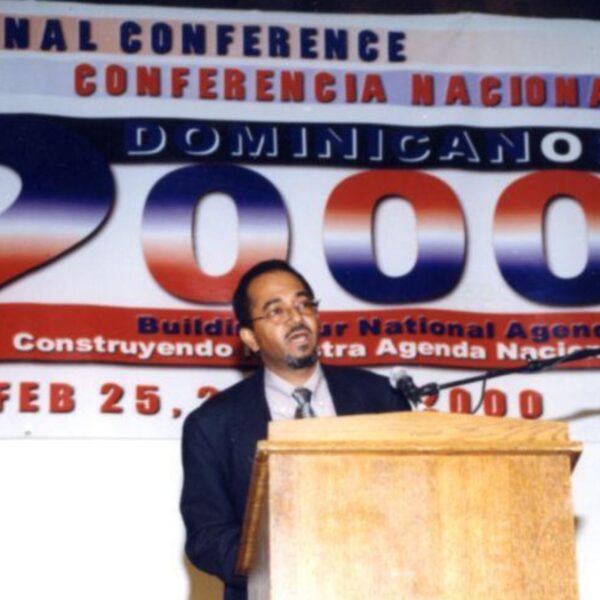 Man speaking at a podium beneath a large banner reading “National Conference Dominicanos 2000: Building Our National Agenda,” dated February 25, 2000.