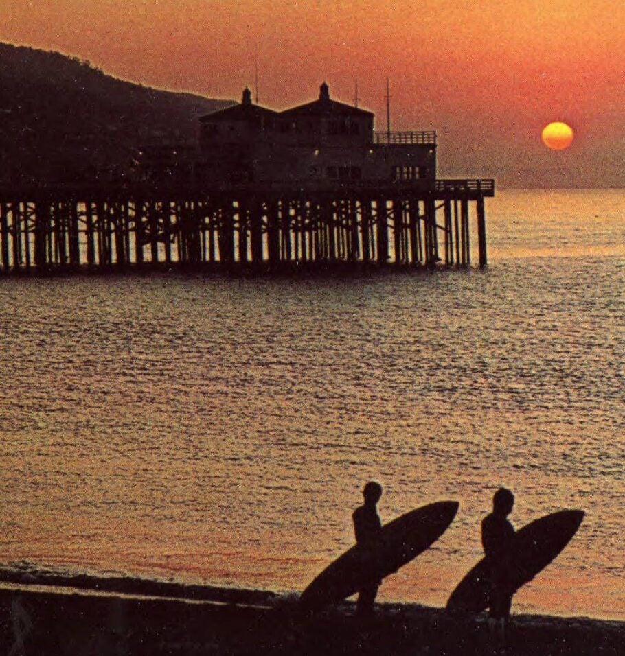 Color postcard-style image of two silhouetted surfers carrying surfboards along the shore at sunrise, with a wooden pier extending into the ocean in the background.