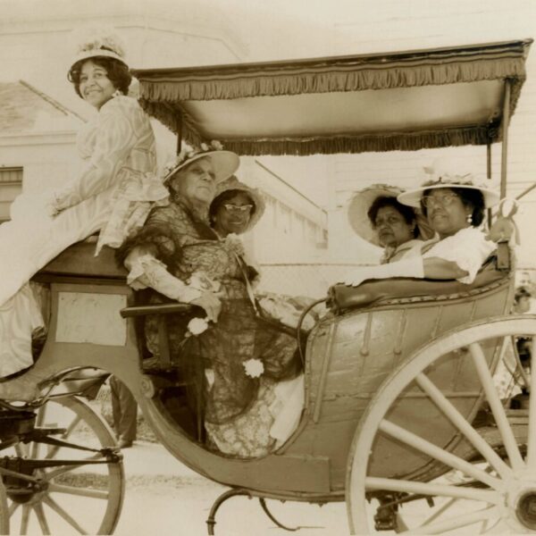 Sepia-toned photograph of five women wearing ornate dresses and wide-brimmed hats, seated in a horse-drawn carriage during a parade or public event.