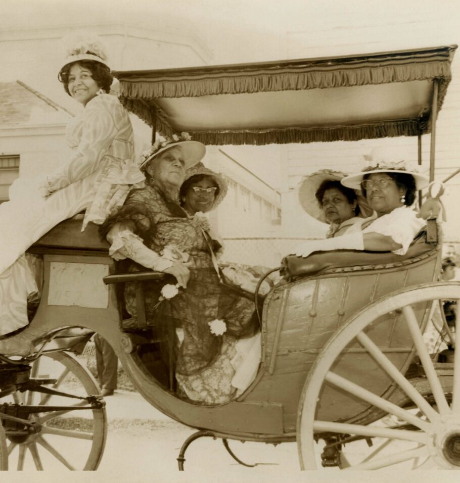 Sepia-toned photograph of five women wearing ornate dresses and wide-brimmed hats, seated in a horse-drawn carriage during a parade or public event.