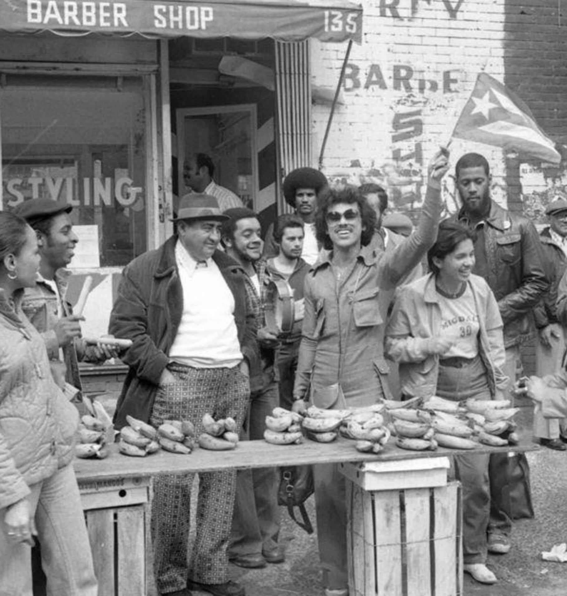 Black-and-white photo of a group of people gathered around a table outdoors, one person holding a Puerto Rican flag, in front of a neighborhood barbershop during a 1971 student strike.