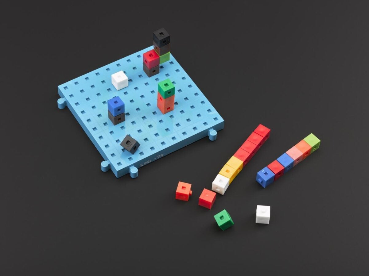 Light blue plastic board with a grid of small square holes, holding multicolored interlocking cubes arranged in short stacks. Additional red, yellow, blue, green, and white cubes are lined up and scattered beside the board against a dark background.