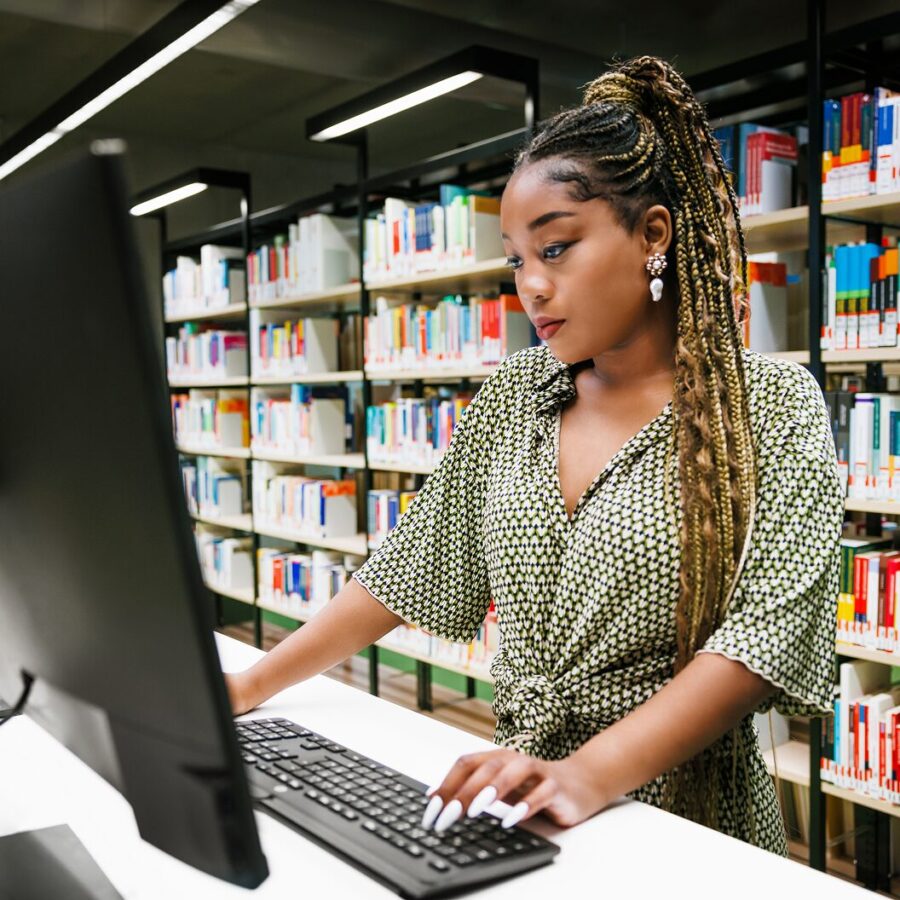 Student using a desktop computer in a library, with bookshelves visible in the background.