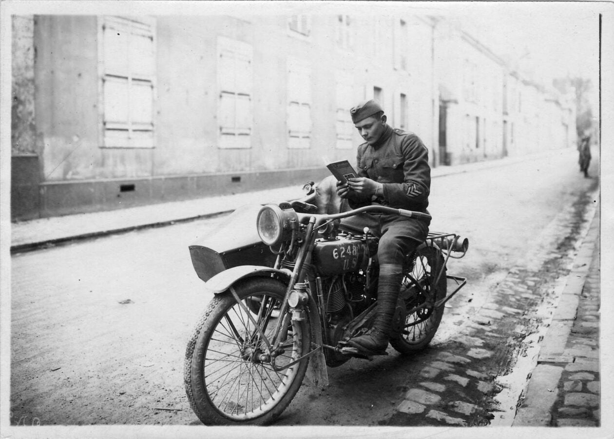 Black-and-white photograph of a uniformed soldier sitting on a military motorcycle on a quiet street, reading a small booklet, with long rows of buildings stretching into the background.