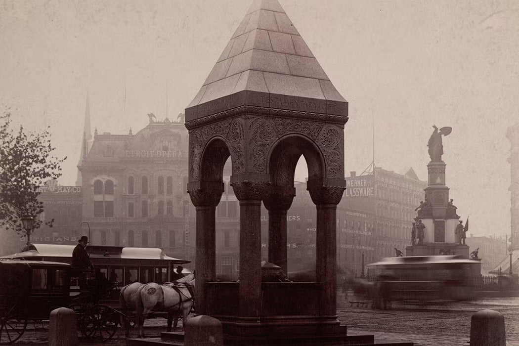 Alt text:
Sepia-toned photograph of the Bagley Fountain in Detroit, featuring a carved stone canopy supported by four columns. A horse-drawn carriage waits nearby, and a blurred streetcar passes in the background. Behind the fountain, late 19th-century buildings and a monument rise through a hazy urban atmosphere.