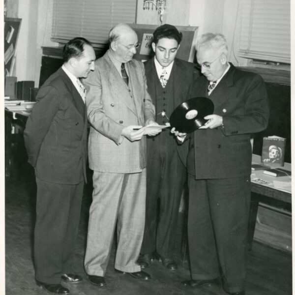 Black-and-white photograph of four men in suits examining a vinyl record and papers in an indoor setting.