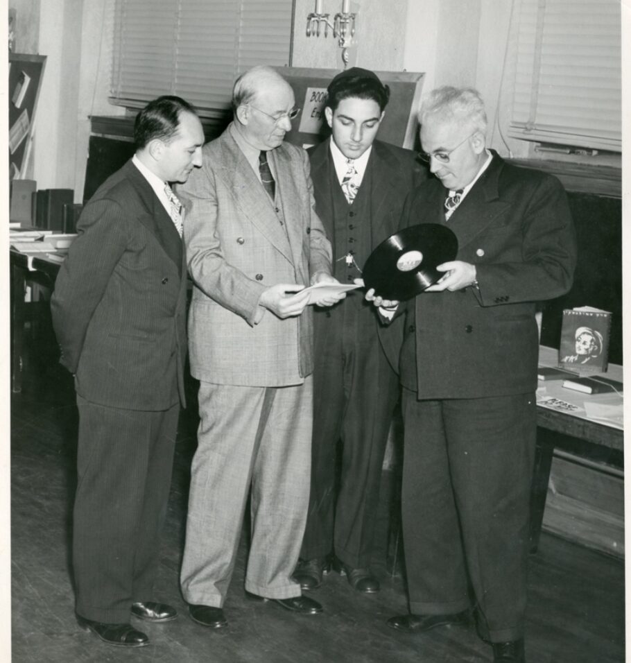 Black-and-white photograph of four men in suits examining a vinyl record and papers in an indoor setting.