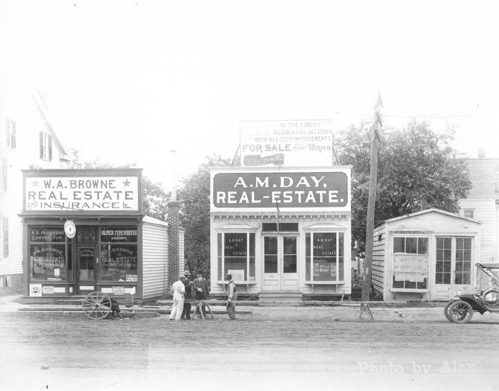 Black-and-white photograph of a small-town street with three storefronts. Two businesses display large signs reading “W.A. Browne Real Estate and Insurance” and “A.M. Day Real Estate.” Several men stand and sit on the sidewalk in front, and an early automobile is parked at the right. Wooden buildings and a dirt road suggest an early 20th-century setting.