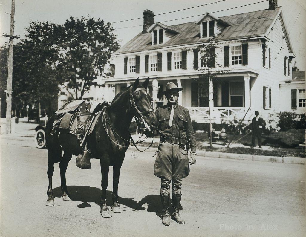 Black-and-white photograph of a military procession moving down a residential street. Uniformed men march in formation carrying American flags and banners, followed by others, including a horse-drawn carriage. An early automobile is parked at the left, and houses line the street, suggesting a World War I–era funeral or commemorative parade.