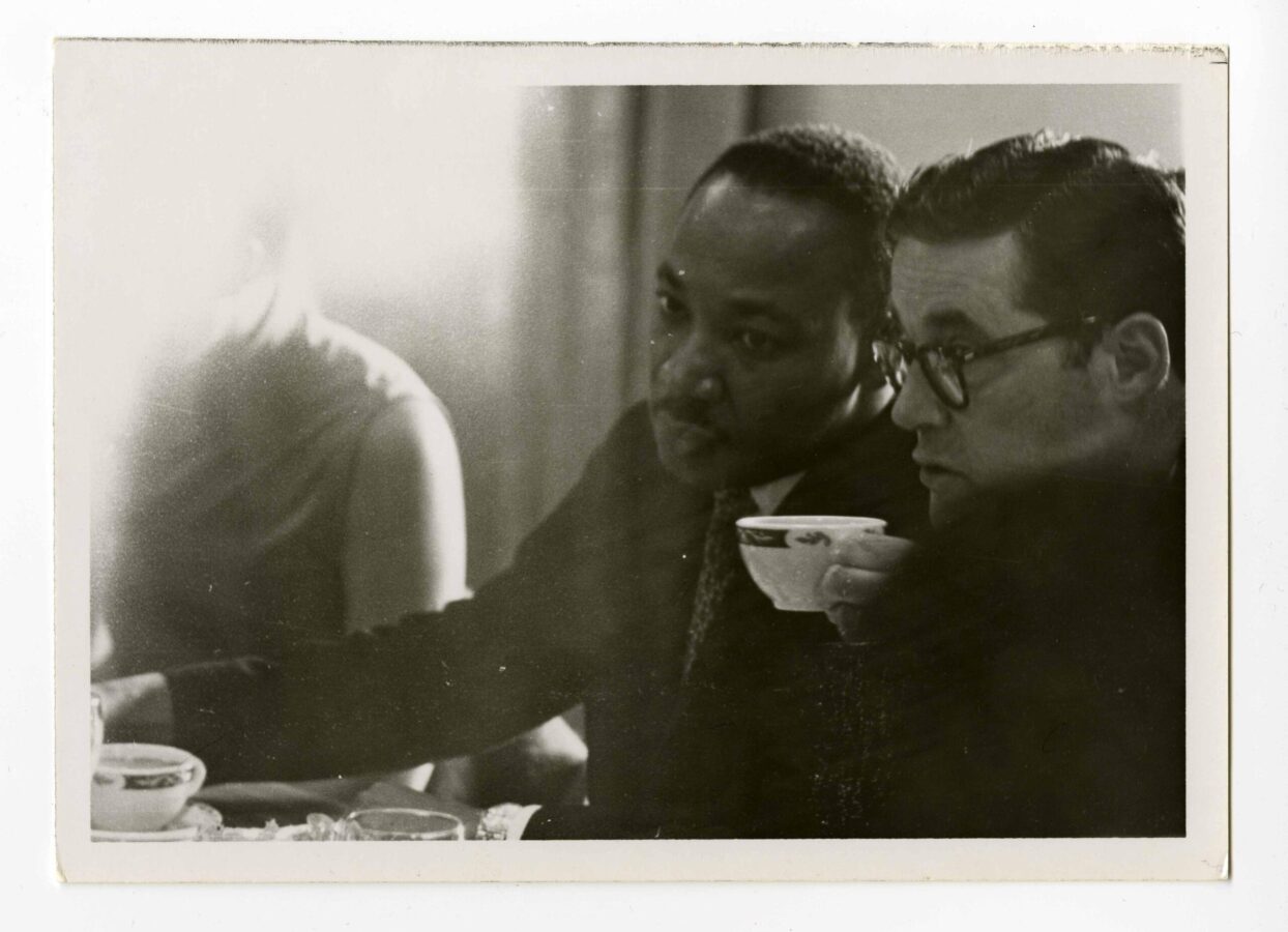 Black-and-white photograph of two men seated closely at a table, leaning forward and looking intently at something off-camera. The man in the center is Martin Luther King Jr., wearing a suit and tie, his expression focused. Beside him, another man wearing glasses holds a teacup near his mouth. A third figure is partially visible on the left, blurred by bright light. Cups and dishes sit on the table in the foreground, suggesting a quiet, candid moment during a meeting or conversation.