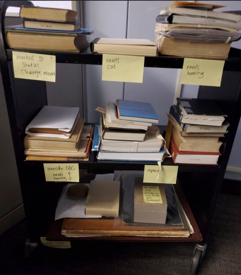 A shelving unit filled with stacks of books and archival materials is labeled with sticky notes indicating processing needs, such as “needs cut,” “needs housing,” “transfer DDC needs housing,” “replace?” and “marked IP status change needed.” The setup visually represents a manual, in-progress archival workflow with items sorted into different categories.