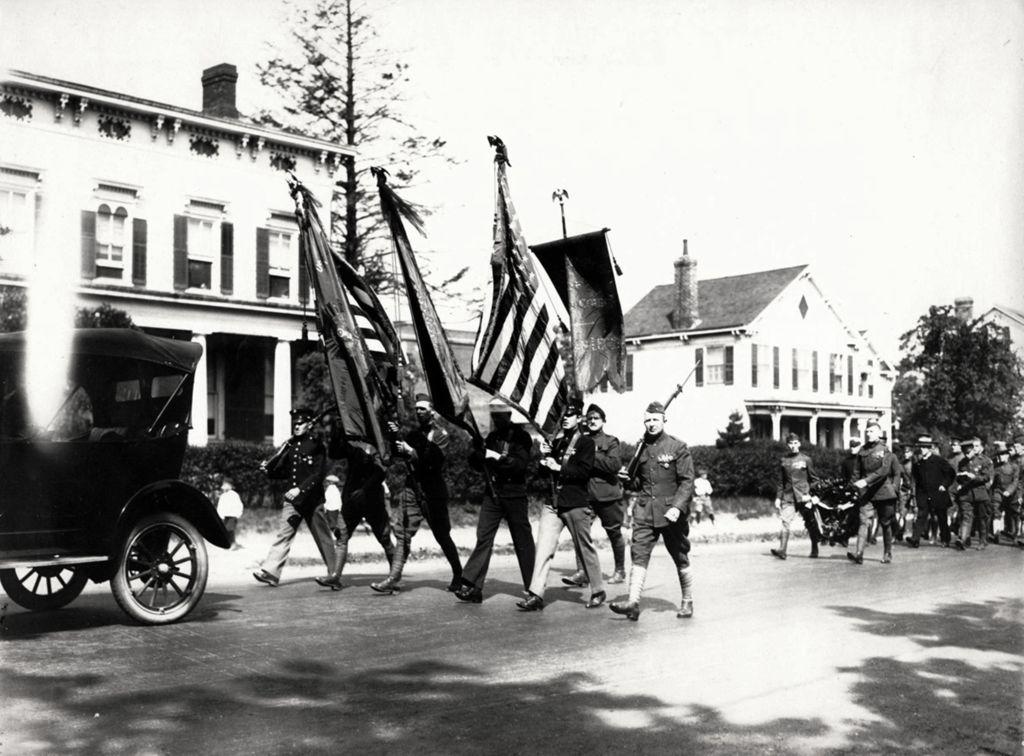 Black-and-white photograph of a military procession moving down a residential street. Uniformed men march in formation carrying American flags and banners, followed by others, including a horse-drawn carriage. An early automobile is parked at the left, and houses line the street, suggesting a World War I–era funeral or commemorative parade.
