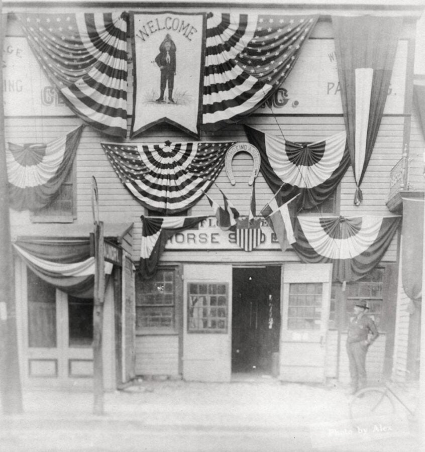 Black-and-white photograph of a storefront heavily decorated with American flags, bunting, and banners. A central sign reads “Welcome” above a figure illustration, and a horseshoe hangs among the decorations. A man stands to the right of the entrance, and the display suggests a patriotic celebration or public event.