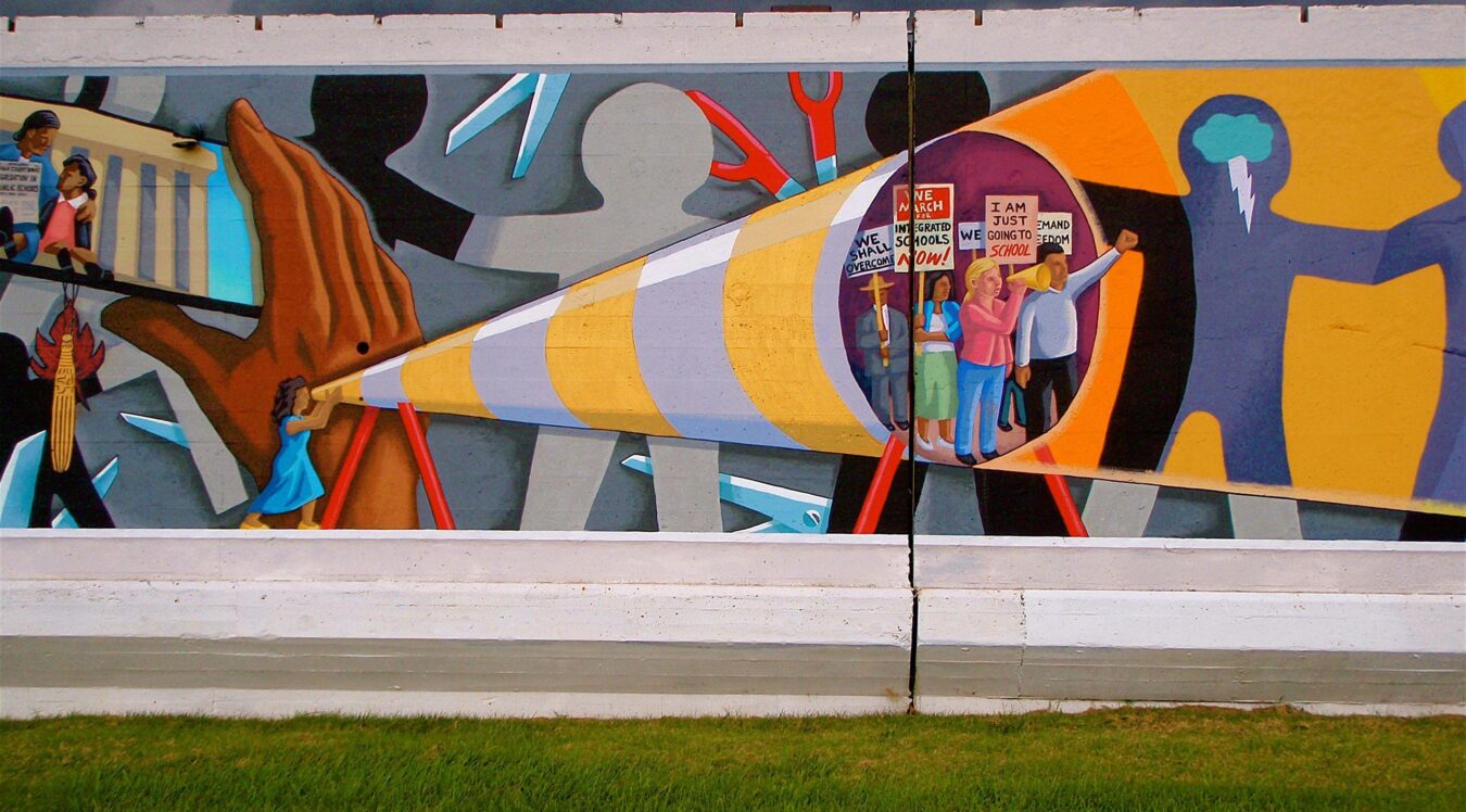 A large outdoor mural painted on a concrete wall depicts a sweeping civil rights and education justice narrative. At the center, a giant pencil rendered in yellow, white, and gray stripes extends diagonally across the composition, held at its base by a small figure in a blue dress. Inside the pencil's hollow tip, a group of diverse protesters carries signs reading "We March for Integrated Schools Now," "I Am Just Going to School," and "We Demand Freedom." To the left, figures march past a columned building , evoking the Lincoln Memorial,  while a corn stalk and fire imagery appear below. To the right, large silhouetted figures in yellow and blue frame the scene, one with a lightning bolt emanating from their head. Scissors, tools, and fragmented shapes fill the background, suggesting barriers being cut through. The overall composition frames education and integration as acts of resistance and power. Sonnet 4.6
