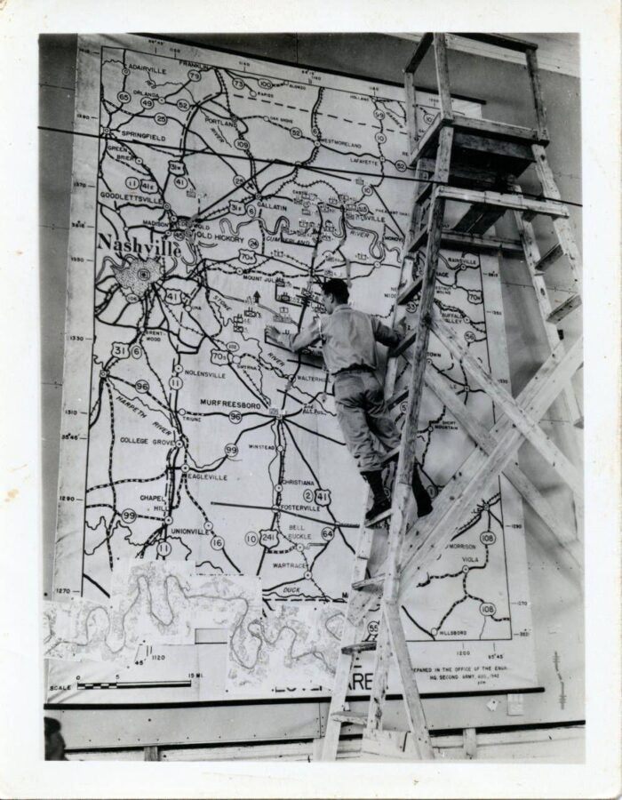 Black-and-white photograph of a man standing on a wooden ladder, working on a large wall-mounted map labeled “Nashville” and surrounding areas. The map shows roads, towns, and routes, and the man appears to be marking or adjusting details near the center. 