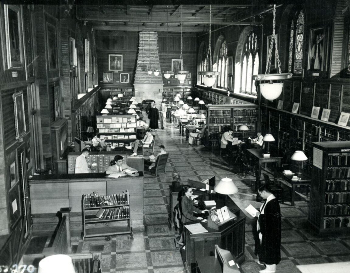 Black-and-white photograph of a large library reading room with bookshelves, study tables, and patrons reading and working beneath high ceilings and hanging lights.