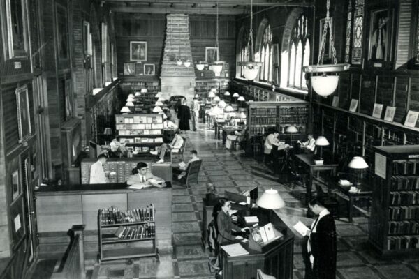 Black-and-white photograph of a large library reading room with bookshelves, study tables, and patrons reading and working beneath high ceilings and hanging lights.