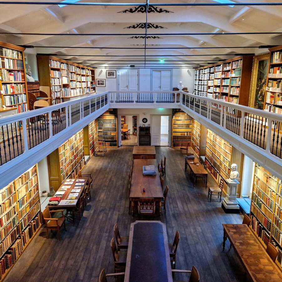 Spacious two-level library with long wooden tables and floor-to-ceiling bookshelves filled with books.