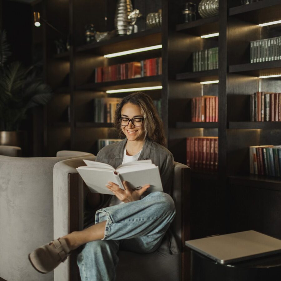 Person sitting in a cozy chair reading a book, smiling, with softly lit bookshelves in the background.