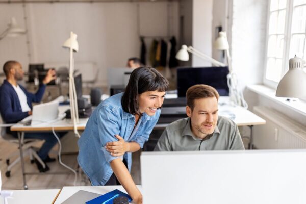 Two colleagues collaborating at a computer in a bright office, smiling while reviewing content on a screen.