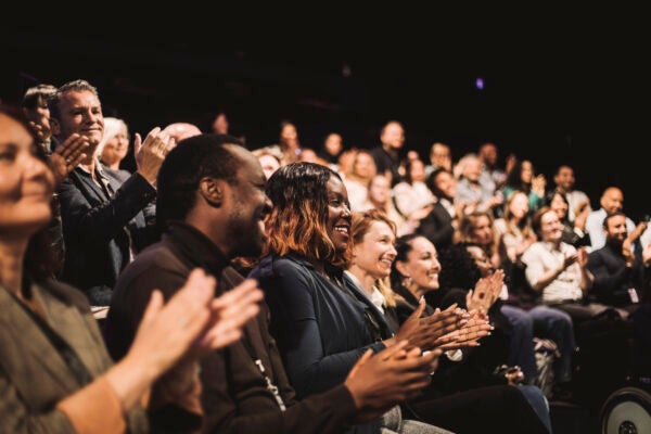 Audience members seated in a dim auditorium applauding during an event or presentation.