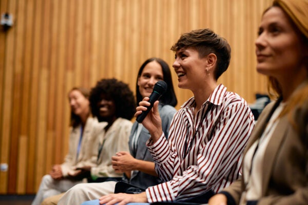 Woman speaking into a handheld microphone during a panel discussion, seated alongside other participants in a wood-paneled conference room.