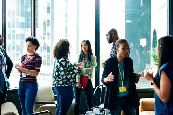 Group of people networking and talking in a bright room with large windows, some holding drinks and wearing event badges.
