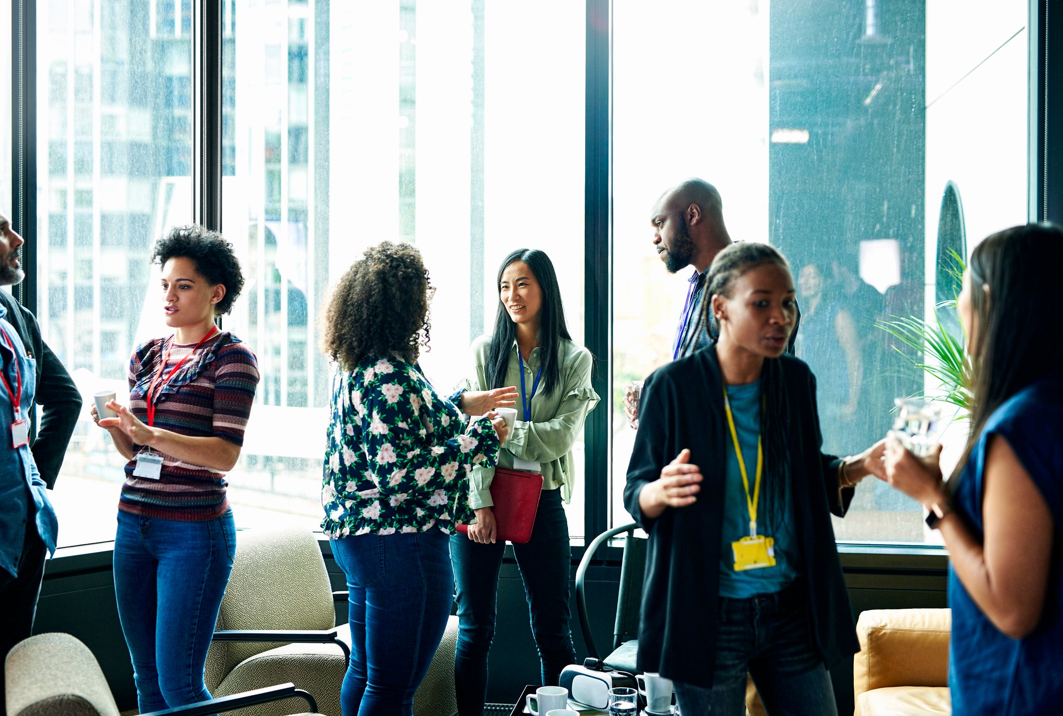 Group of people networking and talking in a bright room with large windows, some holding drinks and wearing event badges.