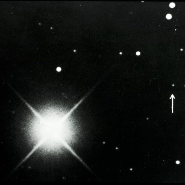Black-and-white astronomical photograph showing a bright star with diffraction spikes on the left and a dense star field; two white arrows on the right point to a small, faint dot identified as Pluto.