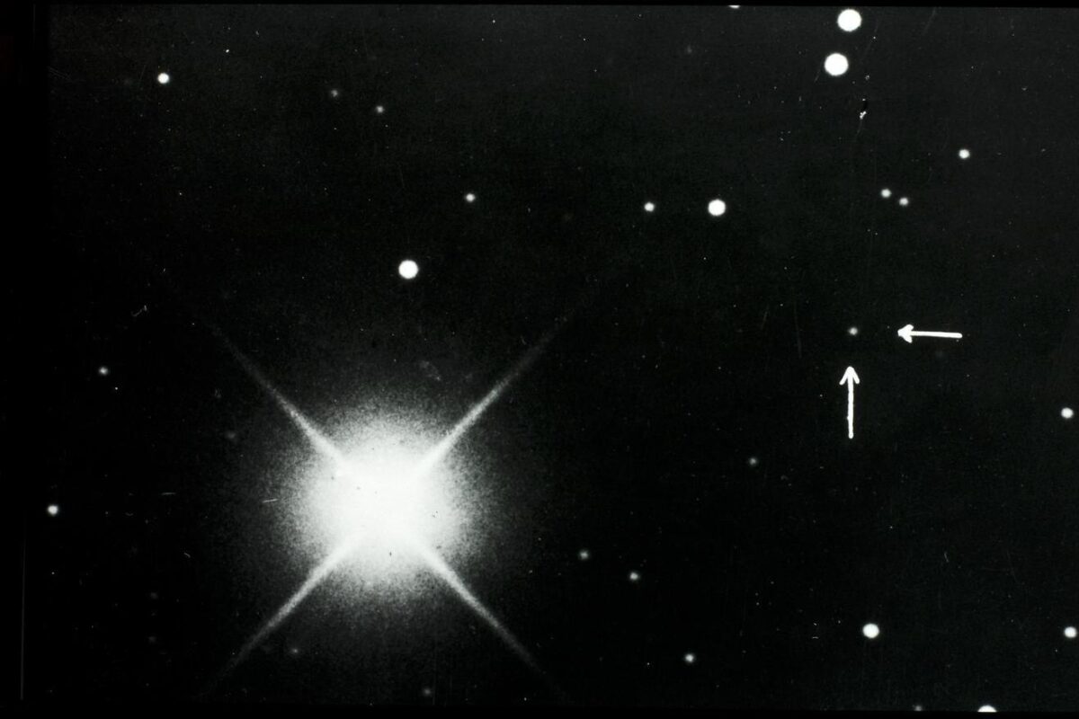 Black-and-white astronomical photograph showing a bright star with diffraction spikes on the left and a dense star field; two white arrows on the right point to a small, faint dot identified as Pluto.