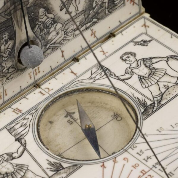 Close-up of an antique ivory diptych sundial and compass designed like a small folding book, featuring engraved Renaissance-style illustrations of figures and scenes, a central magnetic compass with directional markings, and a string gnomon casting a shadow across the hour scale.