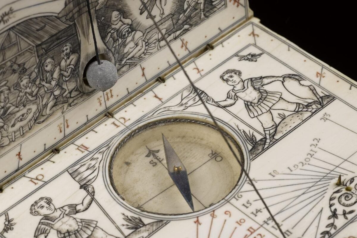 Close-up of an antique ivory diptych sundial and compass designed like a small folding book, featuring engraved Renaissance-style illustrations of figures and scenes, a central magnetic compass with directional markings, and a string gnomon casting a shadow across the hour scale.