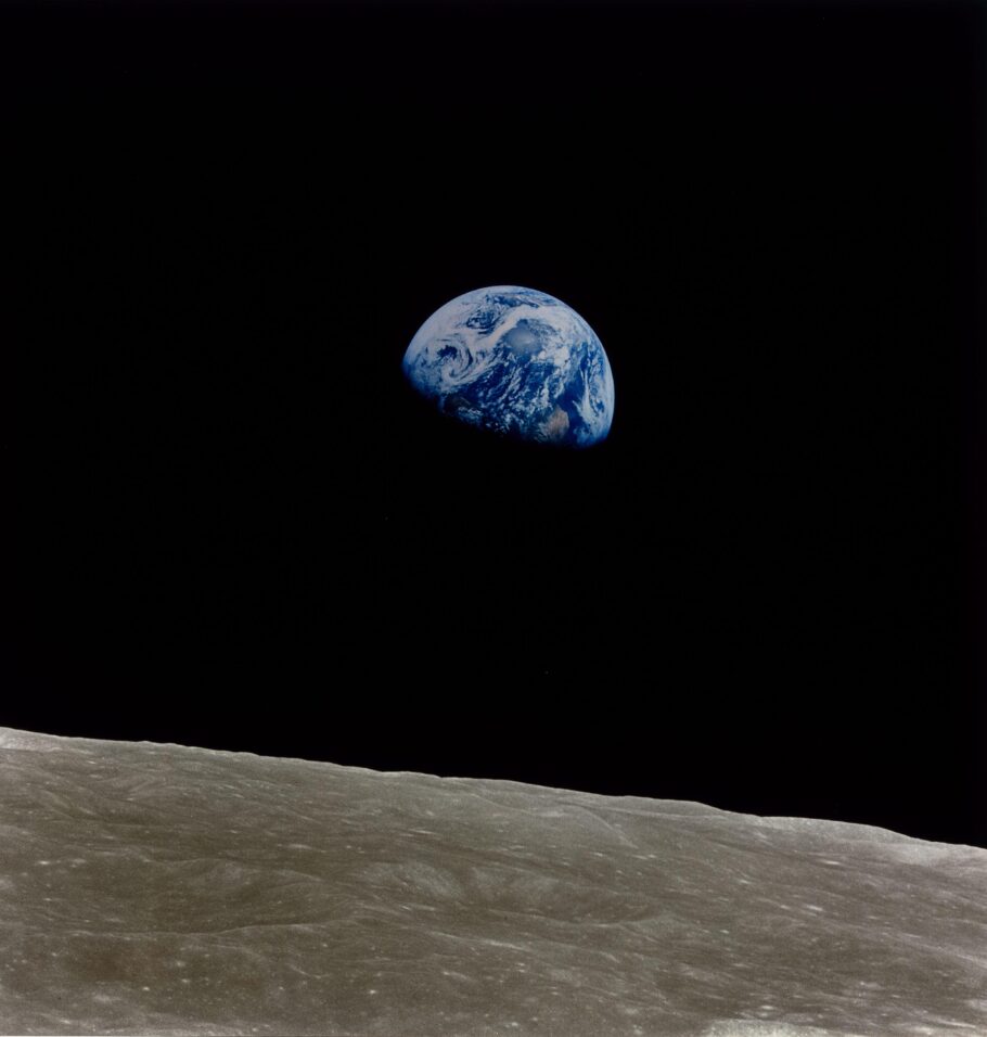 “Earthrise” photograph showing the blue-and-white Earth emerging above the gray, cratered surface of the Moon against the blackness of space.