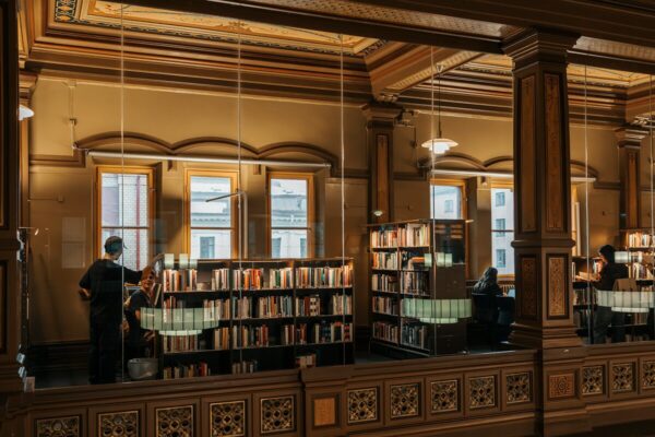 Ornate library interior with tall wooden columns, large windows, and bookshelves; a few people browse and read in a warm, historic setting.