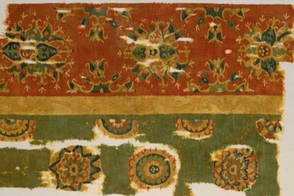 Fragment of a woven tapestry with geometric and floral medallion patterns in rust red, olive green, gold, cream, and black. The upper section features repeating symmetrical motifs on a red ground, separated by a narrow gold band from a green lower section decorated with circular floral designs. The textile is worn and fragmented, with visible tears and missing sections along the edges and center.