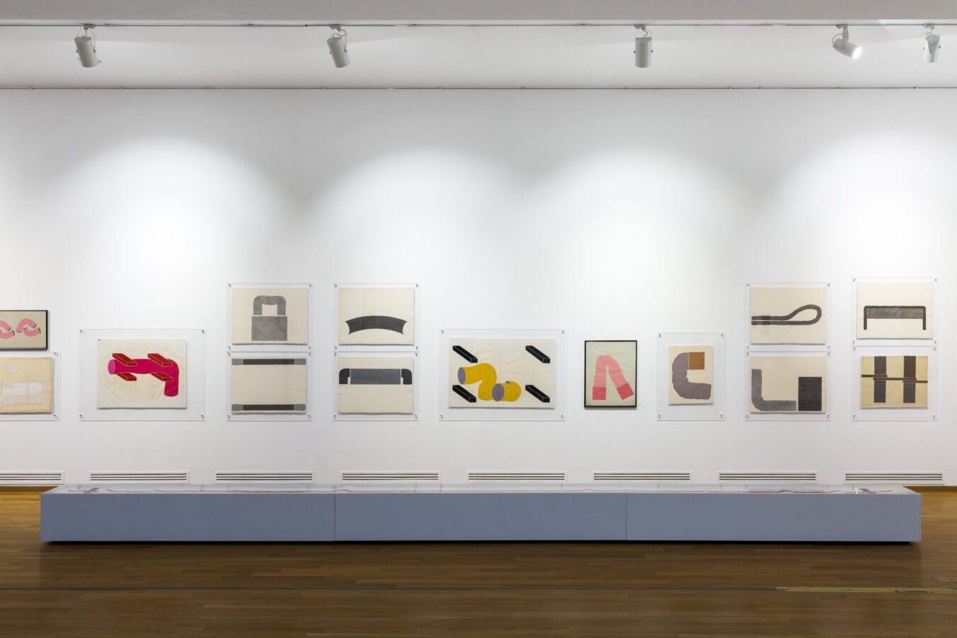 Gallery wall displaying a series of abstract geometric works on paper by Ion Bitzan, arranged in a grid above a long glass-topped display case.