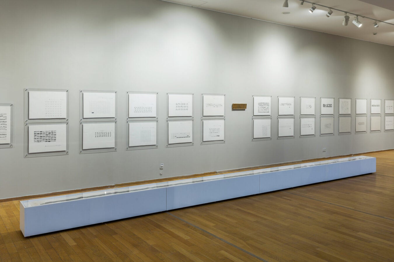 Installation view of minimalist graphic works on paper mounted in transparent frames along a gallery wall, with a glass display case in the foreground.