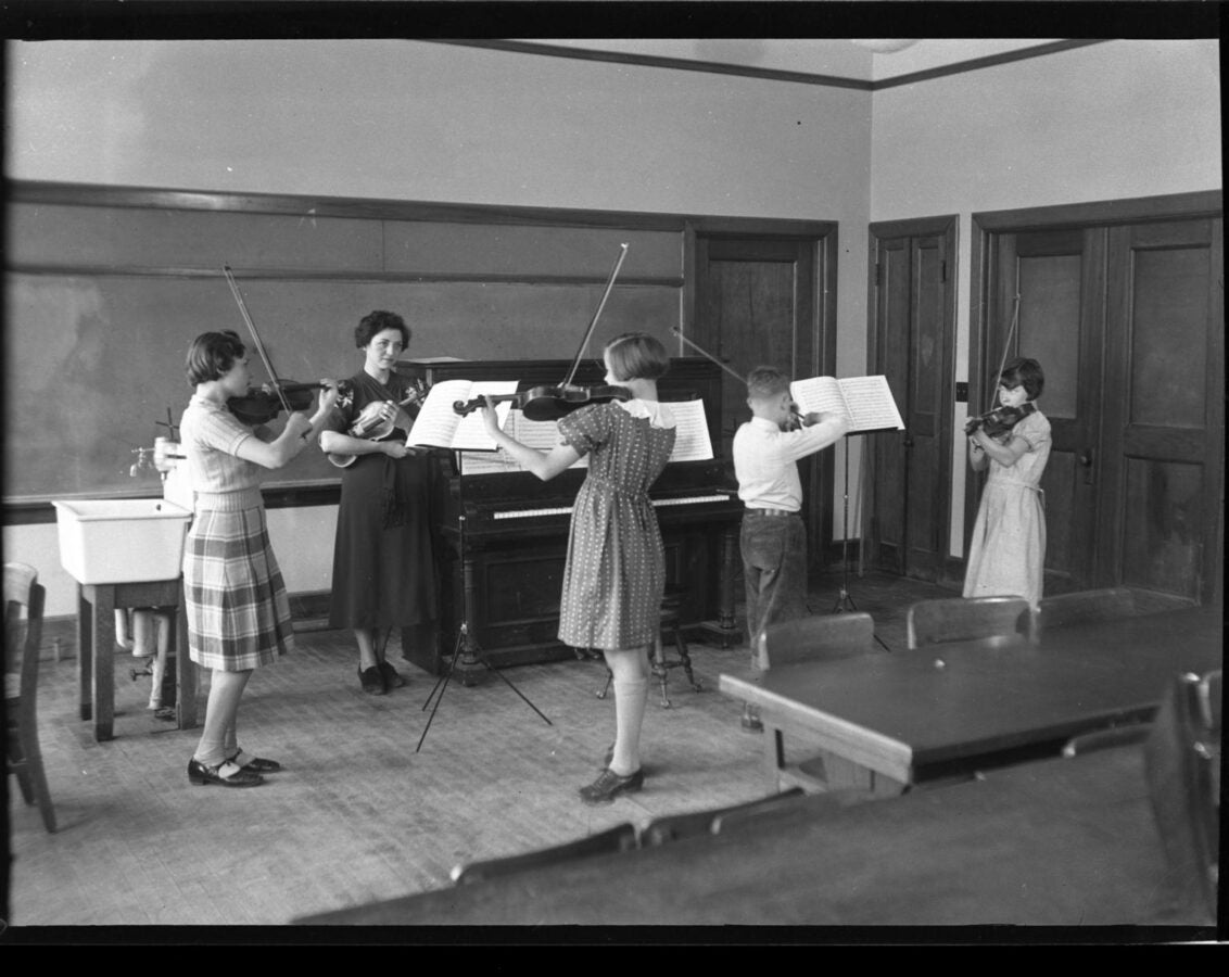 Black-and-white photograph of a music class, with an instructor standing beside a piano while four children play violins and read from music stands.