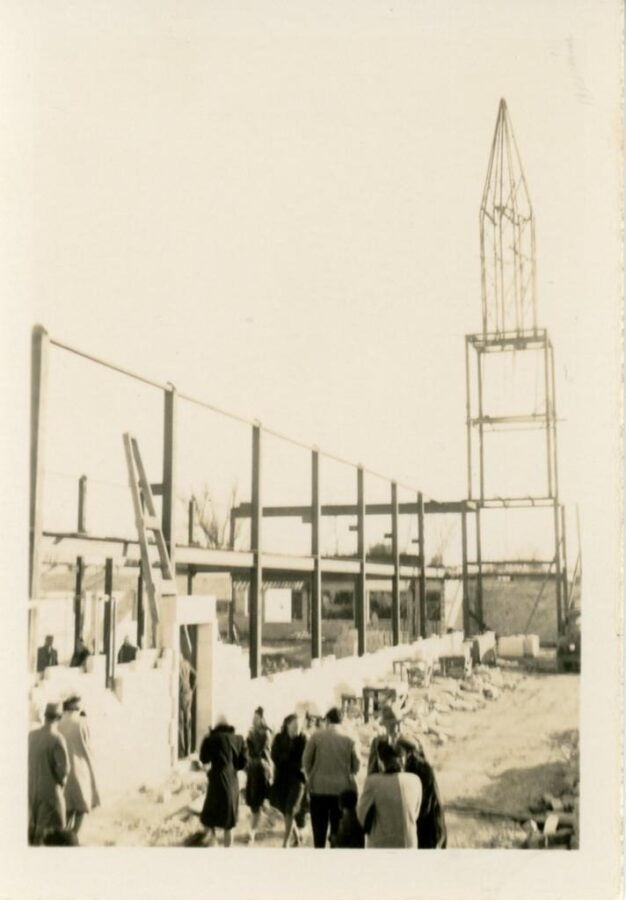 Black-and-white photograph of a steel-frame building under construction, with workers and structural supports visible.