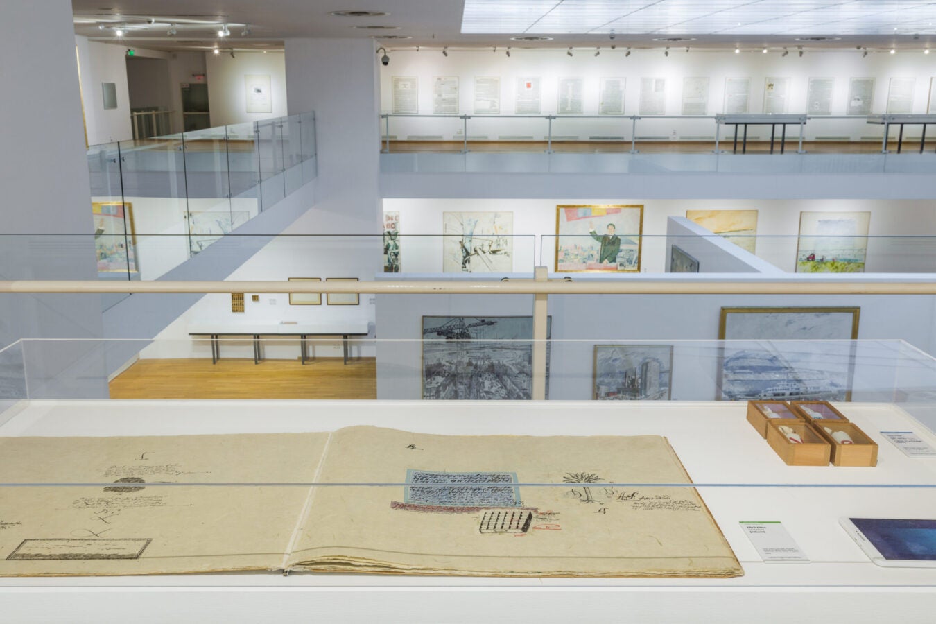Wide view of a bright, modern art museum gallery with multiple levels connected by glass railings. In the foreground, a large open artist’s book or manuscript is displayed in a glass case alongside small boxed objects. Paintings and drawings hang on the walls of the lower level, while framed text-based works line the upper gallery. The space features white walls, wood floors, and overhead track lighting.