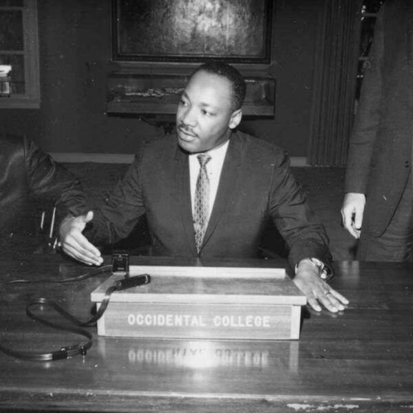 Black-and-white photograph of Martin Luther King Jr. gesturing and speaking while seated at a table with an 'Occidental College' sign.