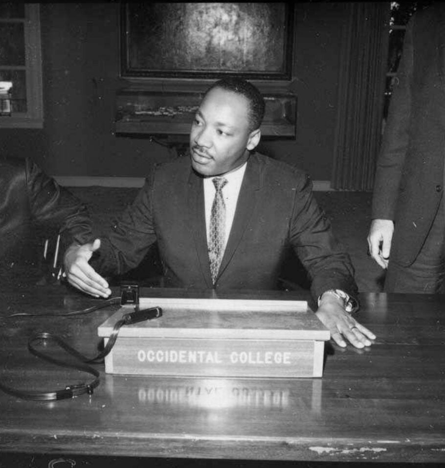 Black-and-white photograph of Martin Luther King Jr. gesturing and speaking while seated at a table with an 'Occidental College' sign.
