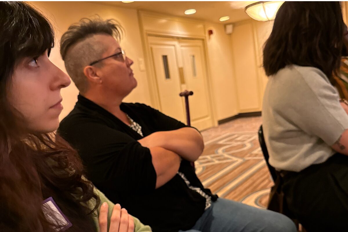 Three conference attendees seated in a hotel meeting space, listening to a speaker outside the frame.