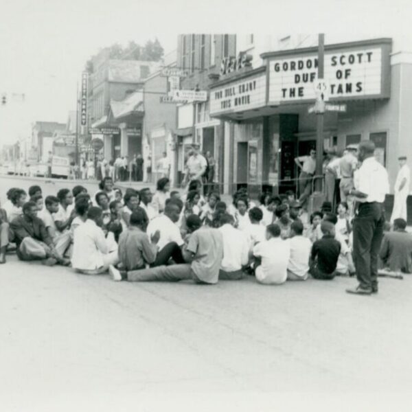 Black-and-white photograph of protesters seated in the street outside a movie theater while onlookers stand nearby.