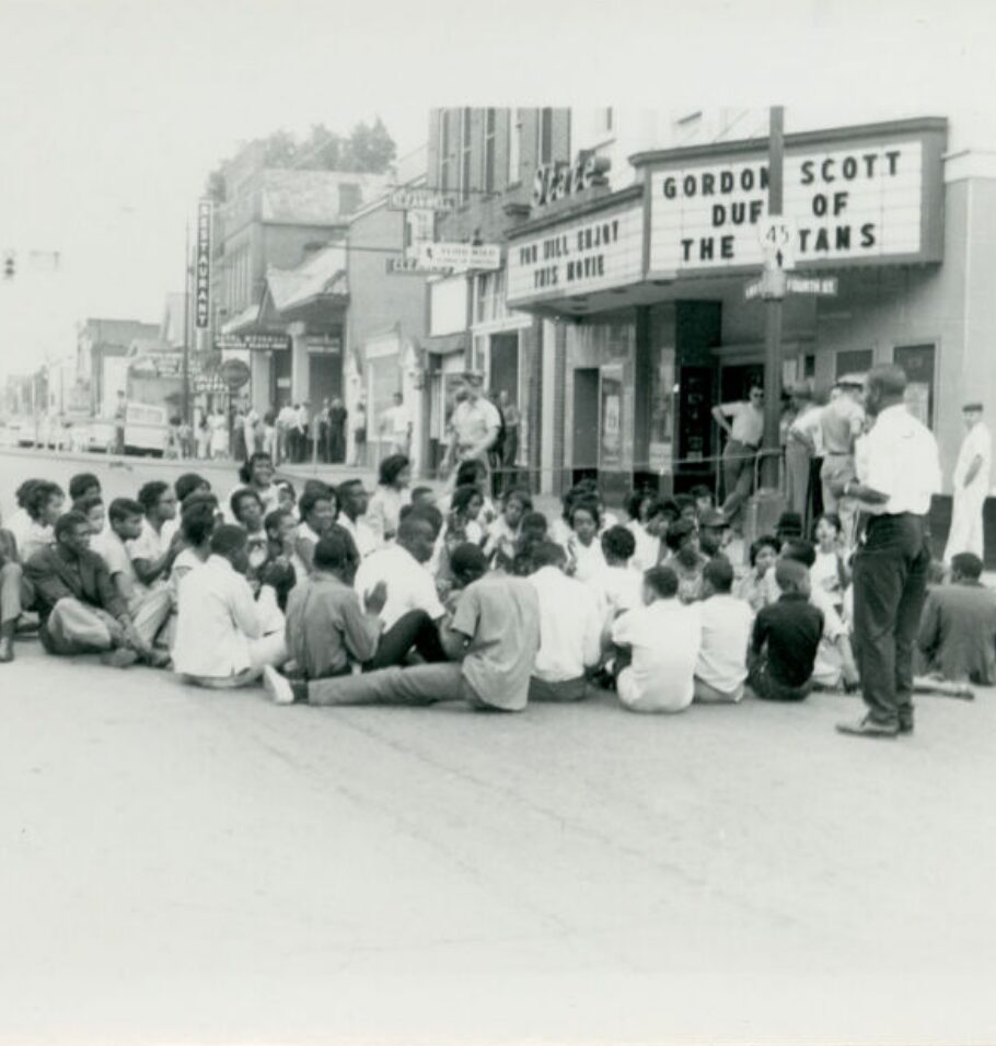 Black-and-white photograph of protesters seated in the street outside a movie theater while onlookers stand nearby.
