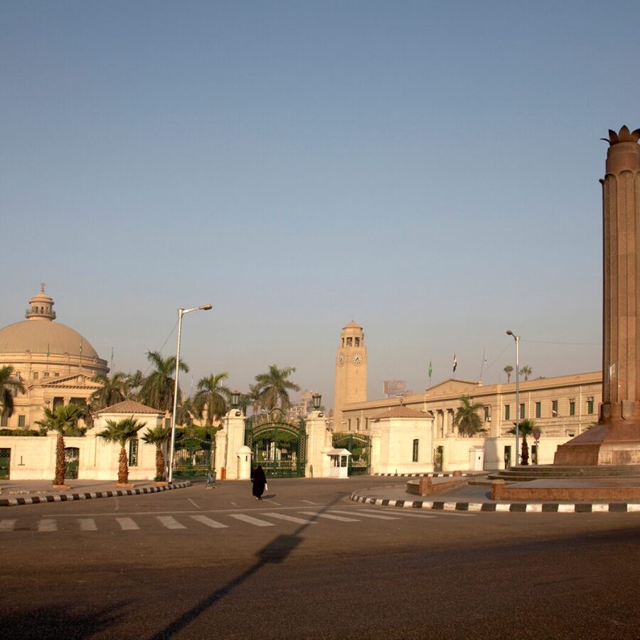 Wide view of a historic university entrance with domed buildings, a clock tower, palm trees, and a tall monument under a clear sky.